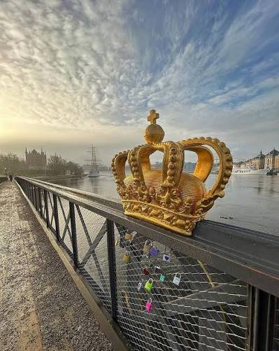 South-western footway of Skeppsholmsbron beside the gilded crown near the middle of the bridge. Stay on the footway, leave passing room and do not lean over the railing.