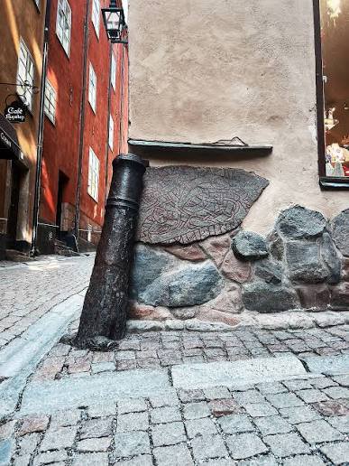 Public pavement at the Prästgatan–Kåkbrinken corner, facing the runestone built into the wall. Keep the narrow passage clear.