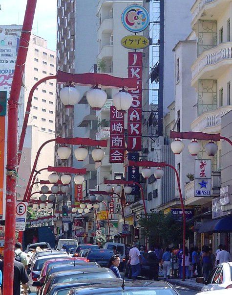 Public pavement at Praça da Liberdade where Rua Galvão Bueno begins, beside a red ornamental streetlamp and clear of crossings.