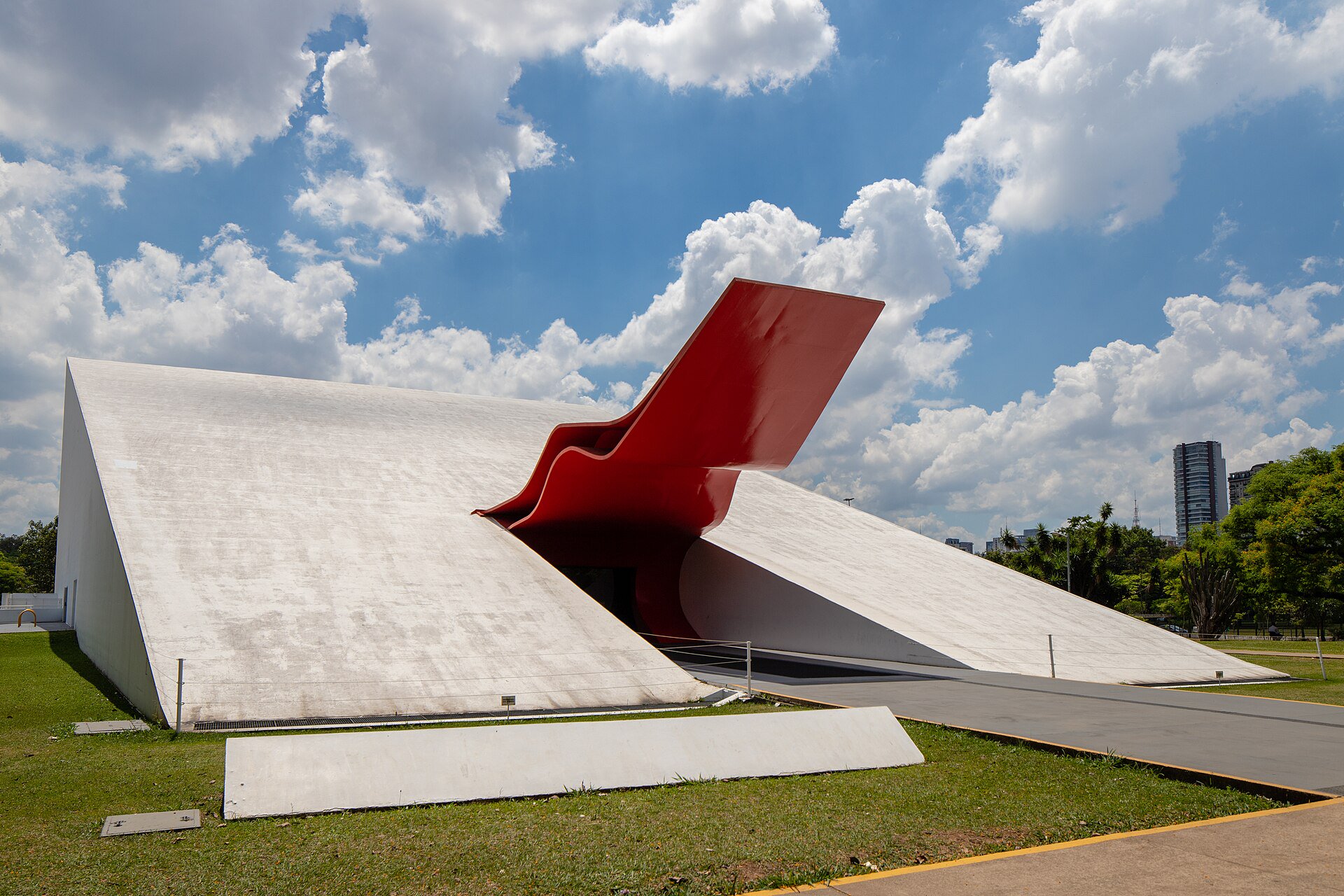 Open park path facing the red entrance canopy of Auditório Ibirapuera, inside Parque Ibirapuera and away from event queues.